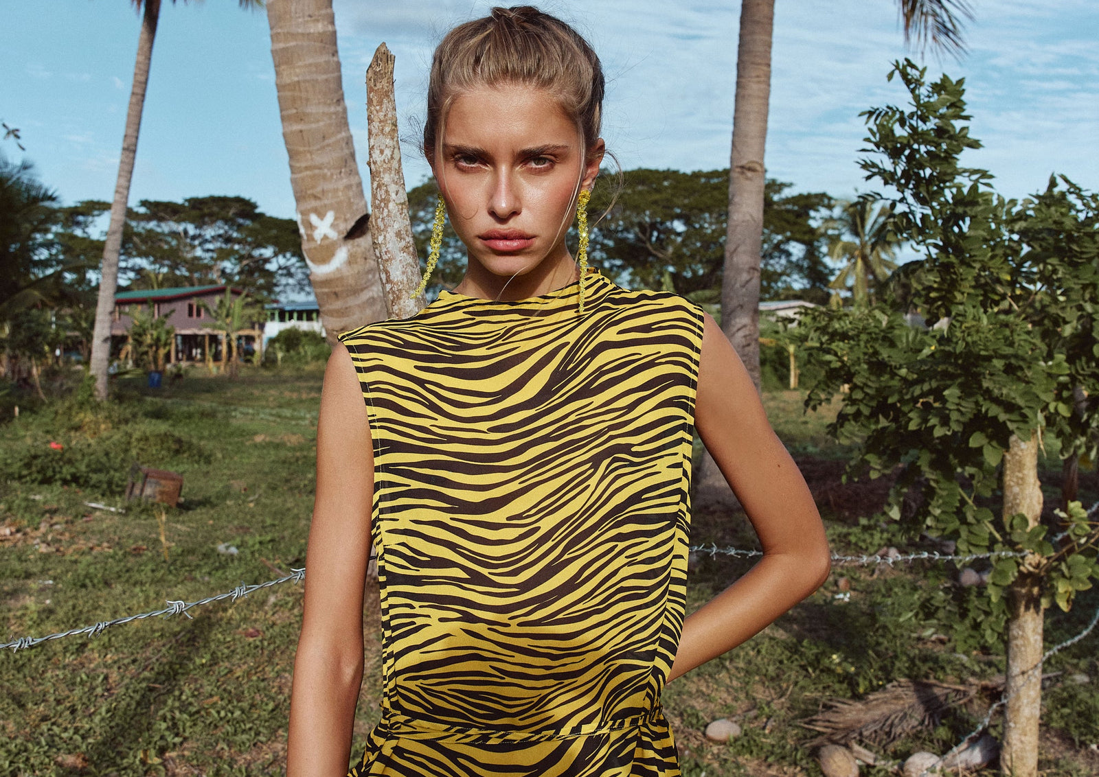 A model stands outdoors in a tropical setting with palm trees, wearing a sleeveless yellow and black tiger-striped top paired with a white and coral skirt.